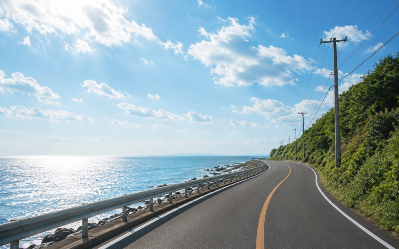 青空の下、穏やかな海沿いを緩やかに続く道路。これからも歩み続ける未来をイメージした風景。